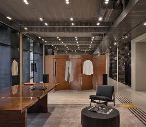 Modern industrial-style mens clothing boutique interior with polished walnut table, dark grey armchair, concrete ceiling, and clothing display racks.
