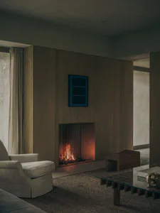 Modern living room interior with a built-in fireplace set within a walnut wood wall panel, a beige swivel armchair, a sculptural wooden stool, and a glass coffee table on a textured rug.
