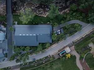 Aerial photograph of a modern rectangular house with dark standing seam metal roof, surrounded by landscaped garden, next to a rocky river and paved driveway with parked cars.