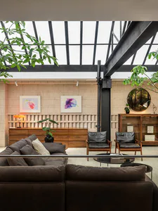 Modern living room with large brown sectional sofa in foreground, two black leather armchairs, wooden credenza, floral watercolor art, and a large glass skylight overhead with visible steel beams and indoor trees.