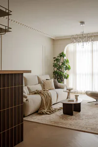 Modern living room featuring a cream sectional sofa, walnut wood paneling, large crystal chandelier, beige area rug, and potted tree by an arched window with sheer curtains.