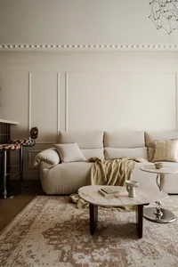 Modern neutral living room with a large off-white sectional sofa, round marble coffee table, patterned area rug, and decorative crown molding on the wall.