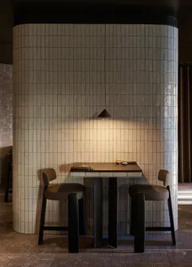 Intimate high-top dining table for two set against a curved wall covered in glossy beige zellige tiles, with two dark wood bar stools and a triangular pendant lamp casting warm light.