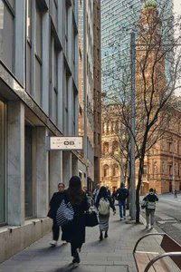 View down a busy city sidewalk flanked by a sleek grey marble building on the left and historic golden sandstone structures on the right, with pedestrians walking away from the camera under natural daylight.