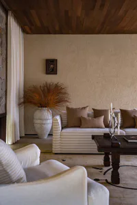 Modern rustic living room interior with white and brown striped sofa, dark wooden coffee table, large white ceramic vase with dried grasses, white armchair in foreground, and textured beige walls under a wood-paneled ceiling.