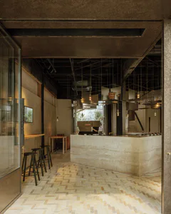 View into a modern industrial cafe with a large travertine stone counter, dark metal ceiling, geometric pendant lights, and light herringbone patterned floor tiles.
