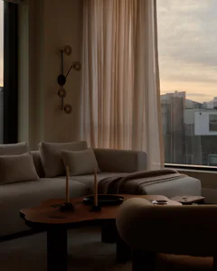 A contemporary living room featuring a light beige boucle sectional sofa with matching pillows, a dark wood irregularly shaped coffee table holding two tall candles, a modern black wall sconce with multiple circular lights, and a large window revealing a city skyline at sunset with sheer curtains.