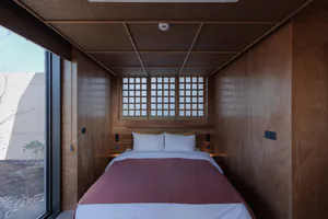 Narrow modern Japanese-style bedroom featuring warm wood paneling, a low platform bed with white and terracotta bedding, shoji-style grid windows, and a large glass door opening to an outdoor garden area.
