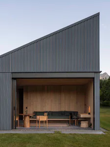 Modern cabin with dark corrugated corten steel exterior and open indoor-outdoor living area furnished with a green sofa, wooden chairs, and a round wooden table, set against a mountain backdrop.