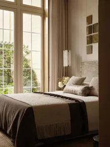 A serene contemporary bedroom featuring a bed with textured beige and brown linens, a woven headboard, wall sconce lighting, and large windows overlooking a lush garden.