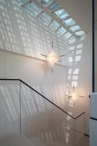 Modern minimalist staircase with glass railings, white walls, and two illuminated starburst pendant lights hanging beneath a long skylight that casts geometric shadow patterns.