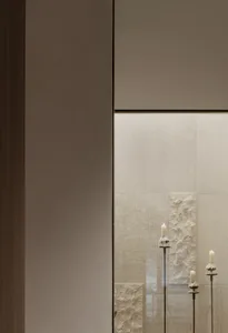 A minimalist interior corner featuring a smooth taupe wall on the left and a textured off-white limestone wall on the right, with three lit white candles in elegant black metal holders of varying heights.