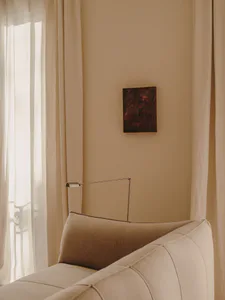 Close-up of a beige linen sofa with a modern articulated floor lamp and a dark abstract painting on a warm beige wall, next to sheer white curtains.