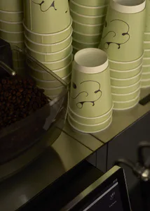 Close-up of sage green paper coffee cups with black abstract face designs stacked on a counter next to a bin of coffee beans in a cafe setting.