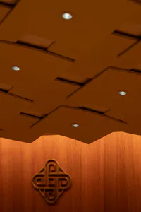 View looking up at a modern coffered ceiling with recessed spotlights and a large golden Chinese knot symbol mounted on a warm wood-paneled wall.