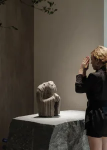 A woman with blonde hair in a black outfit stands with hands pressed together in prayer, facing a weathered headless stone Buddha statue displayed on a large granite pedestal in a minimalist interior space with neutral walls and soft lighting.