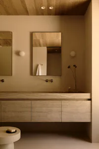 Modern minimalist bathroom with a long travertine floating vanity, rectangular mirror, spherical wall sconces, wooden ceiling, and a small round side table with soap.