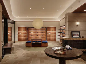 Interior view of a high-end shoe store with wooden shelving units displaying various shoes, a large spherical pendant lamp, blue upholstered seating, a round wooden table with trophies and decor, and a stone fireplace with framed sports photograph.