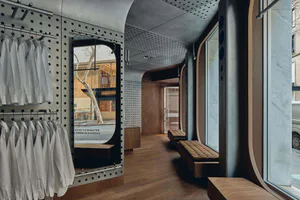 Modern boutique store interior with curved walnut wood walls, built-in upholstered benches, perforated steel clothing racks holding white shirts, and large arched windows overlooking the street.