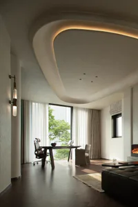 Modern minimalist home office and living room with a dramatic curved plaster ceiling featuring integrated warm lighting, dark wood flooring, large windows with sheer curtains overlooking green trees, a dark wood desk with ergonomic chair, and a fireplace on the right.