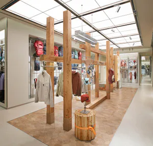 現代零售空間內部，大型回收木材柱梁結構作為服裝陳列系統，展示多款戶外機能外套與背包，搭配石紋地板與天窗天花板。