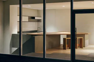 Modern minimalist kitchen viewed through glass doors at night, featuring a large live-edge oak kitchen island with concrete base, floating shelves, and soft recessed lighting.