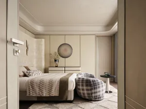 View into a modern luxury bedroom with a neutral color palette, curved headboard, plaid lounge chair, and built-in cabinetry.