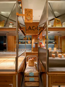Narrow corridor inside a bakery lined with wooden shelves, stainless steel display cases, flour sacks, and a hanging pineapple puff pastry sign.