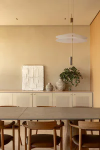 Modern minimalist dining room featuring a long oak dining table with matching wooden chairs, a sculptural white pendant light, a textured plaster artwork, and ceramic vases with eucalyptus on a light wood sideboard against a beige wall.