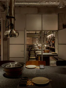 Interior view of an industrial-style Japanese shabu-shabu restaurant with concrete walls, stainless steel exhaust hoods, and set dining counters in warm dim lighting.