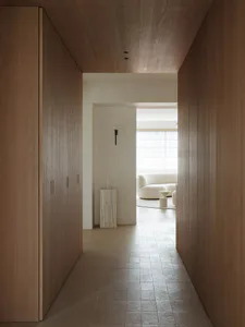 Long narrow hallway with floor-to-ceiling oak wood cabinetry and matching wood ceiling, leading to a bright living room with a curved white sofa and marble pedestal.