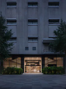 黃昏時分一座粗獷風混凝土建築的正面視角，開放且明亮的現代玻璃門廳入口，兩側有圖案屏風、樹木和低矮景觀，位於鋪面庭院。