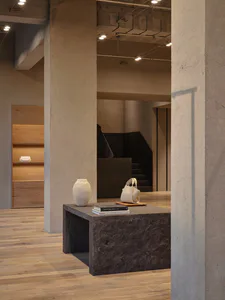 Modern brutalist interior featuring a dark concrete console table with a ceramic vase, white handbag, and stacked books, set against raw concrete walls and light oak flooring.