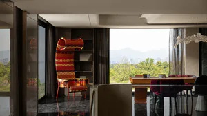 Modern dining room interior featuring a tall high-back armchair with vibrant red, orange and black geometric pattern, wooden dining table with burgundy velvet chairs, large floor-to-ceiling windows overlooking green mountains and cityscape, dark marble floor, and sculptural white chandelier.