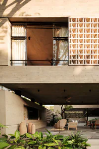 Modern tropical house outdoor courtyard featuring exposed concrete architecture, wooden lattice screen, wicker lounge chairs, and lush tropical plants under warm sunlight.