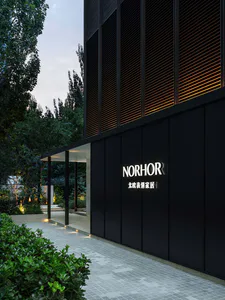 現代黑色建築立面，白色 NORHOR 發光標誌與中文文字，搭配水平木格柵溫暖背光，旁邊是景觀花園步道、樹木與地面燈光，時值黃昏。