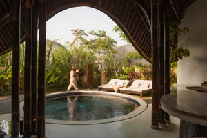 View from inside a dark bamboo pavilion looking out through a large arched opening to a circular swimming pool in a lush tropical garden courtyard, with a staff member carrying a tray and two lounge chairs visible in the background.