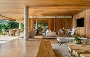 Bright tropical modern living room featuring a warm teak wood ceiling, concrete pillar, stacked stone wall, white sofas, and natural wood furniture opening to a garden.