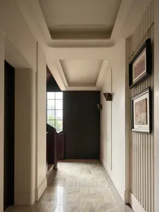 Long elegant interior hallway with light marble herringbone floor, off-white fluted walls, dark wood door, wall sconces, and framed artwork gallery wall.