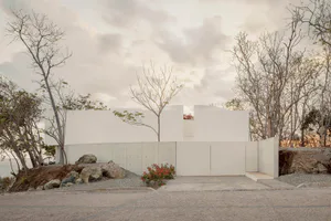 Modern minimalist concrete house with white geometric walls, set on a rocky cliffside surrounded by leafless trees under a cloudy sky, with a paved driveway in the foreground.