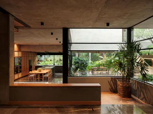 Modern open-plan kitchen and dining area with concrete ceiling, wooden cabinetry, large glass walls overlooking a lush tropical garden and patio.