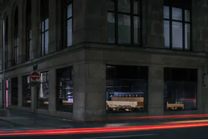 Nighttime street view of a modern minimalist restaurant with concrete facade, large glass windows revealing wooden tables and warm lighting, with red light trails from passing vehicles in the foreground.