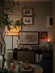 Cozy living room corner featuring a dark wood console table styled with a lit floor lamp, vintage turntable, champagne bottles, framed sheet music, and a flickering candle, with books and a potted tree in the foreground.