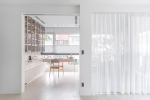 Modern minimalist home office featuring light wood built-in storage, a wooden desk and chair, large windows with roller blinds, and sheer white curtains on the right.