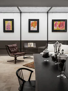 Modern living and dining area with three framed floral watercolor paintings on a white and dark grey wall, a brown leather and wood lounge chair, a black wooden dining table with glass apothecary jars, and a black spindle-back chair in the foreground.