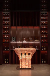 A modern luxury wine cellar featuring a glowing orange onyx bar counter with three oval cutouts, surrounded by dark wood shelving units filled with wine bottles and crystal decanters under warm ambient lighting.