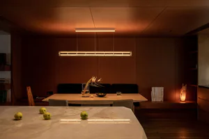 Modern dining room featuring a long cylindrical linear pendant light, walnut wood panel walls, a wooden dining table with dark upholstered bench and grey chairs, and a foreground marble table with green apples under warm ambient lighting.