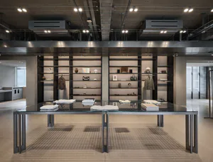 Modern industrial-style luxury clothing boutique interior featuring exposed concrete ceiling, dark steel shelving units, and a large glass-top display table in the foreground.
