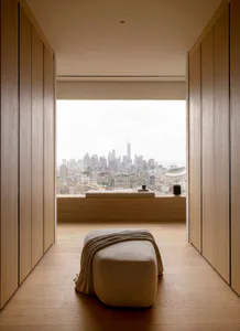 Modern minimalist interior with light oak built-in cabinetry on both sides, a low window seat with cushion, and a large floor-to-ceiling window offering a panoramic view of the New York City skyline including One World Trade Center.