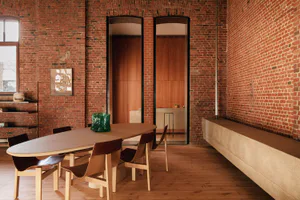 Spacious industrial loft dining area featuring a long oval wooden table, six modern wooden chairs with dark leather seats, exposed red brick walls, and two arched doorways leading to a kitchen with wooden cabinetry.